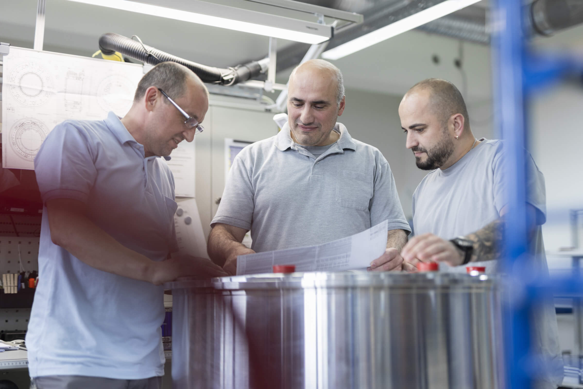Three technicians in a production laboratory analyze technical drawings over a metal cylinder.