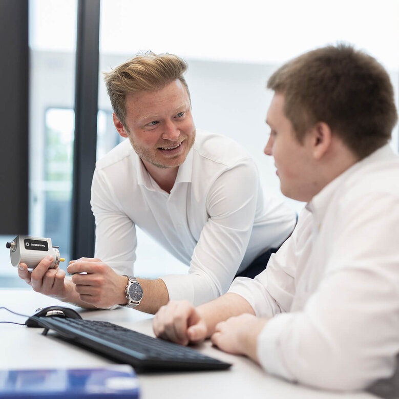 Two men talking in an office, one holding a small industrial camera.