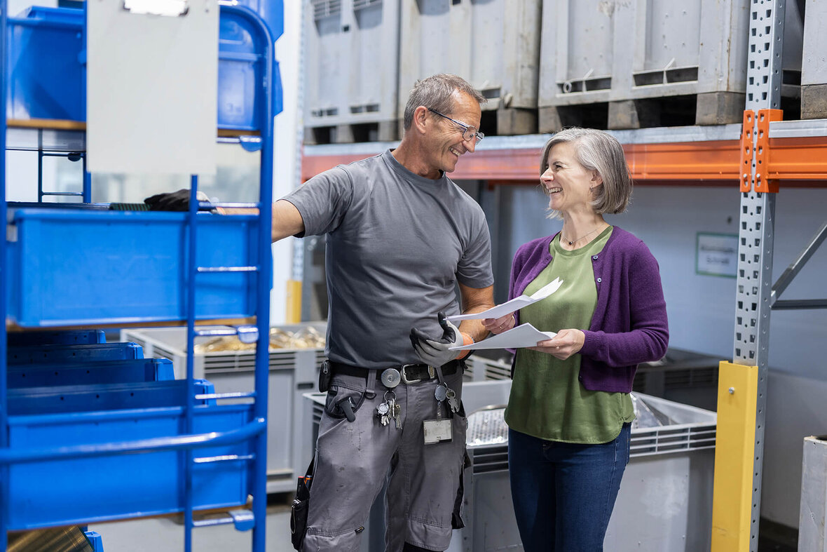 Two colleagues having a friendly conversation in a warehouse while holding documents.