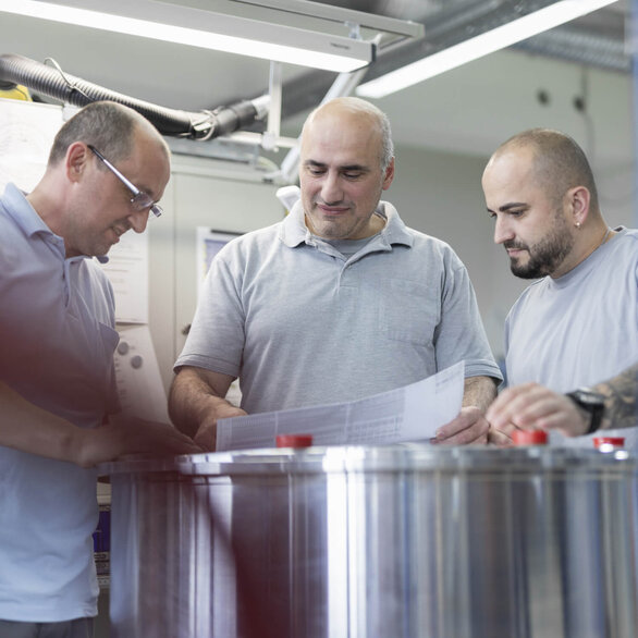 Three technicians in a production laboratory analyze technical drawings over a metal cylinder.
